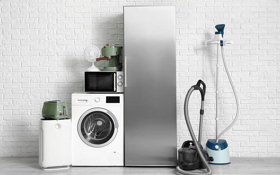 A group of home appliances, including a fridge, washing machine, microwave, and vacuum, against a white brick wall.